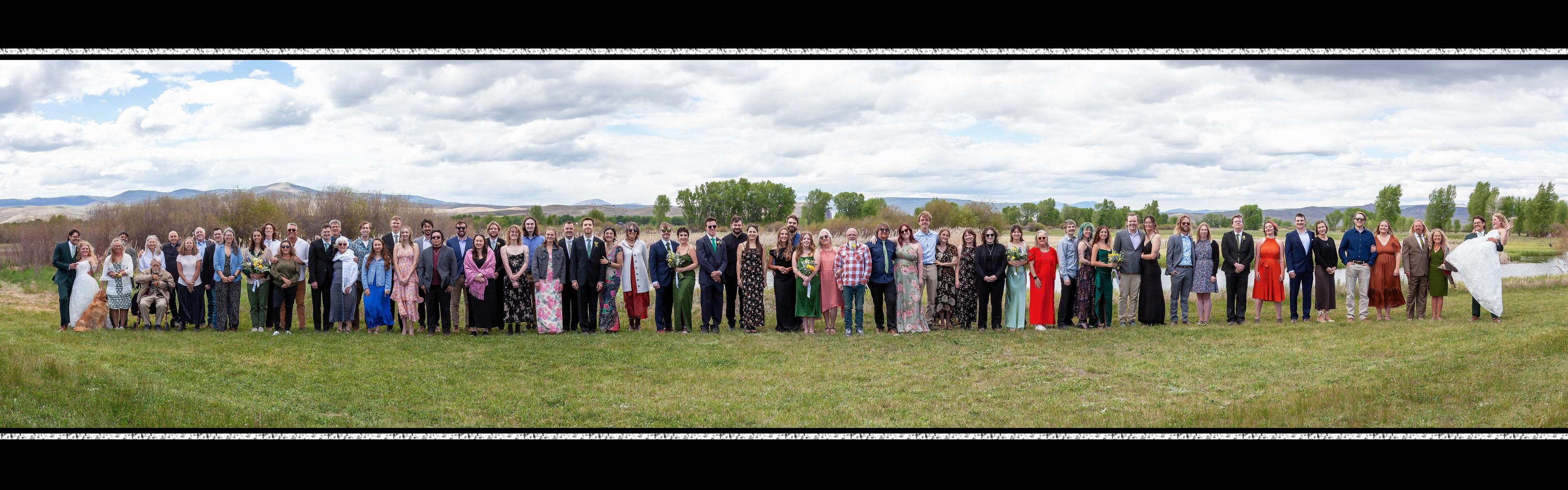 Wedding panoramic photo — large group of guests in formal attire with mountain and meadow backdrop